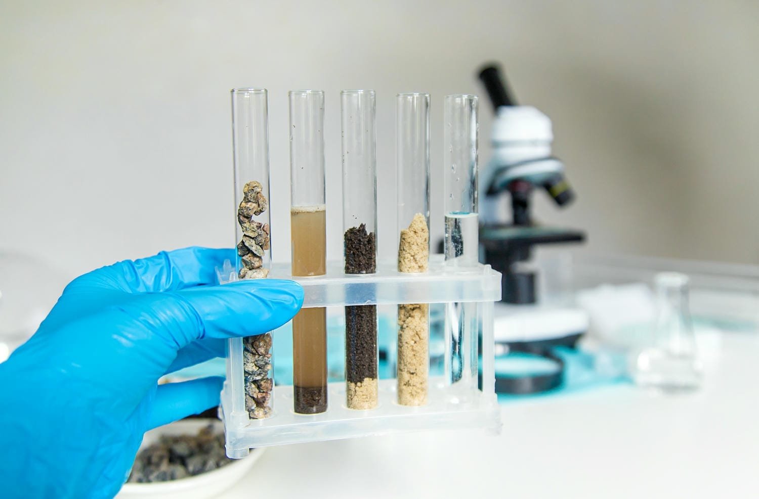 A gloved hand holding test tubes of soil layers beside a microscope